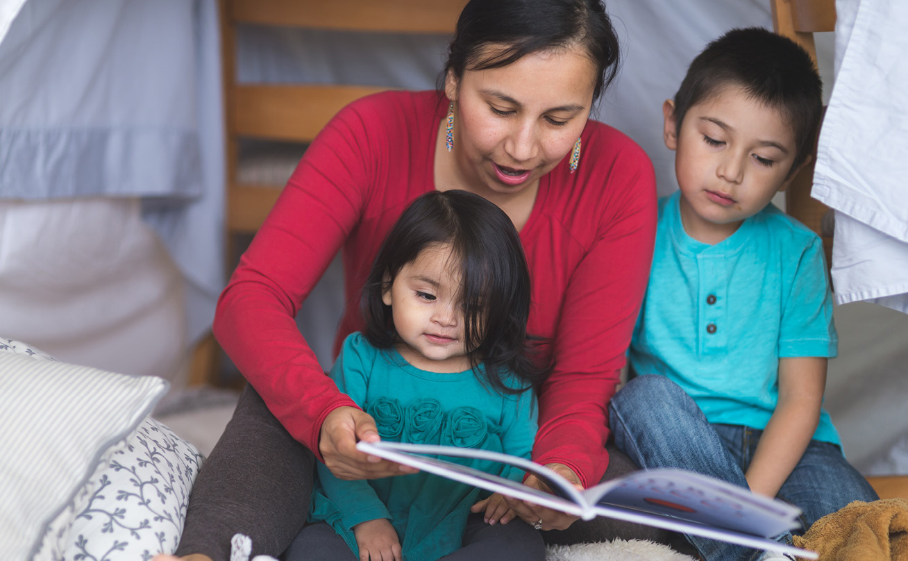 An Indigenous woman reads a book to her two young children.