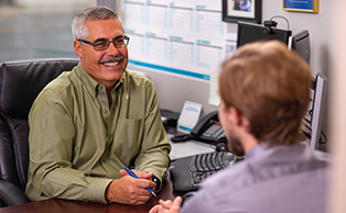 A male employee with glasses and a grey mustache is smiling while meeting with a member.