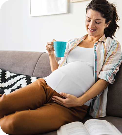 A smiling pregnant woman cradles her belly while sitting on a couch and holding a mug. 