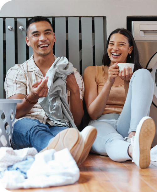A smiling Asian couple sit on the floor next to a washing machine and fold laundry.
